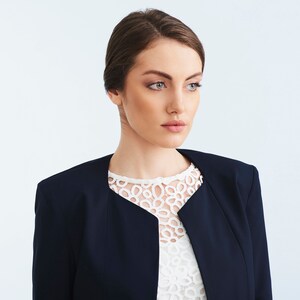 May include: A woman wearing a navy blue blazer over a white lace top. She has brown hair styled in a bun and is looking to the right.