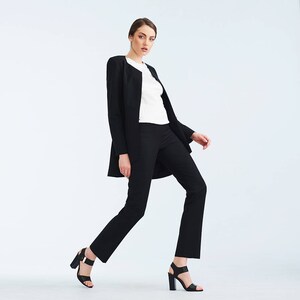 May include: A woman wearing a black blazer and black pants with a white shirt underneath. She is wearing black heeled sandals and is standing with one leg slightly bent.