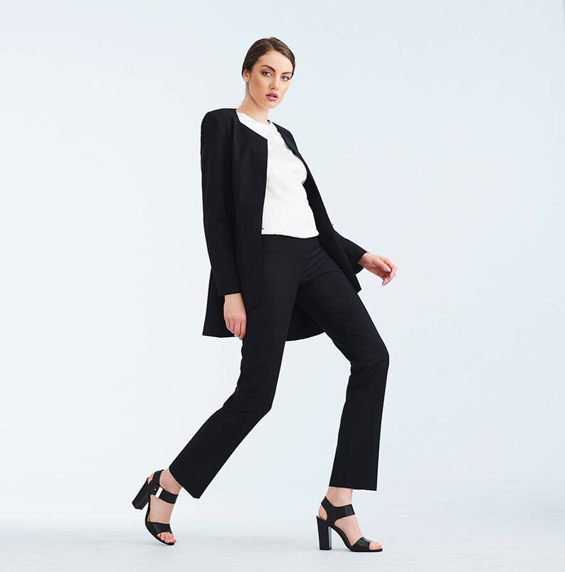 May include: A woman wearing a black blazer and black pants with a white shirt underneath. She is wearing black heeled sandals and is standing with one leg slightly bent.