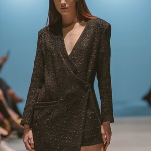 May include: A black wrap dress with a sparkly texture. The dress has a deep V-neckline and long sleeves. The model is wearing the dress with a neutral makeup look and her hair is styled in a straight, long style.