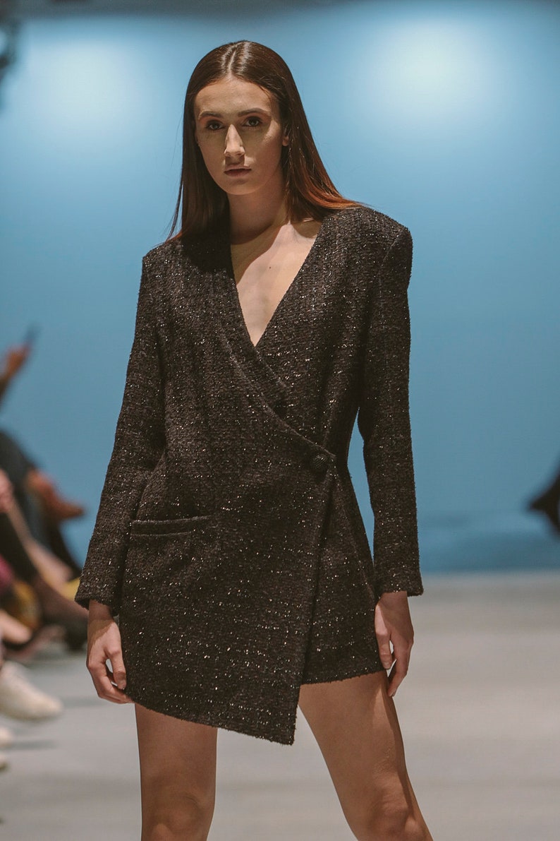 May include: A black wrap dress with a sparkly texture. The dress has a deep V-neckline and long sleeves. The model is wearing the dress with a neutral makeup look and her hair is styled in a straight, long style.