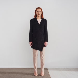 May include: A woman wearing a black tweed blazer with a diagonal closure and a pair of light pink trousers. She is standing in front of a white wall.