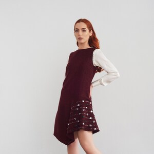 May include: A woman wearing a burgundy dress with a white trim and a white blouse. The dress has a unique asymmetrical hemline.