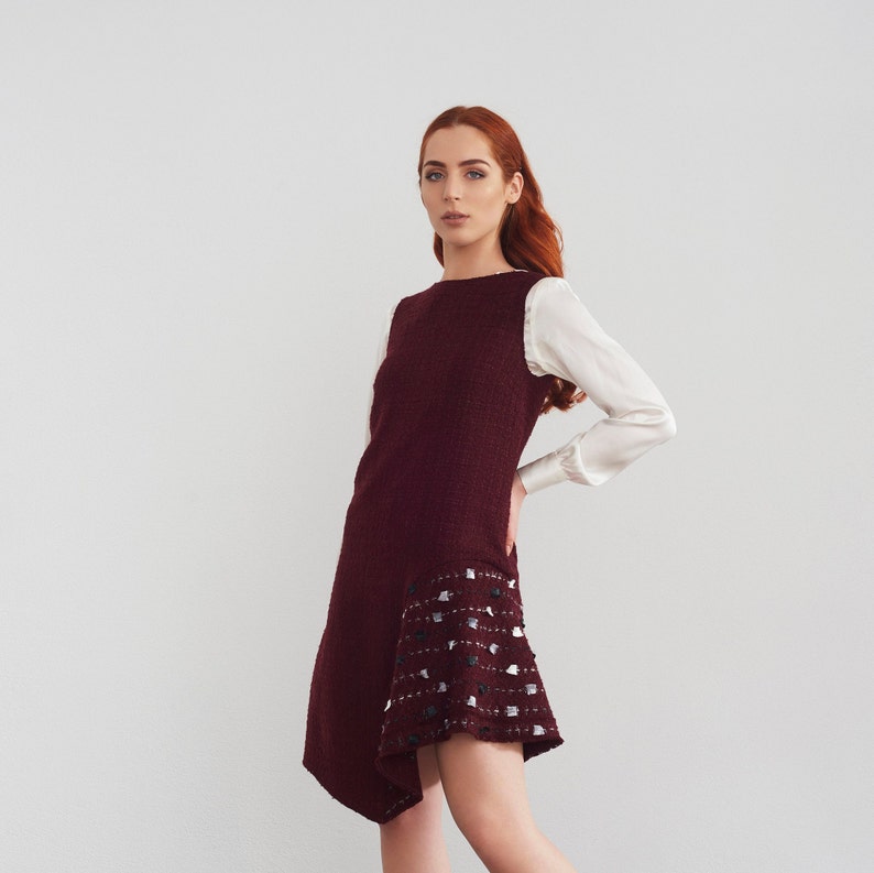 May include: A woman wearing a burgundy dress with a white trim and a white blouse. The dress has a unique asymmetrical hemline.