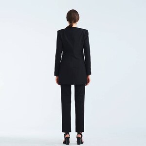May include: A black blazer with a long, fitted silhouette. The blazer has a notched collar and long sleeves. The model is wearing black pants and black heels.
