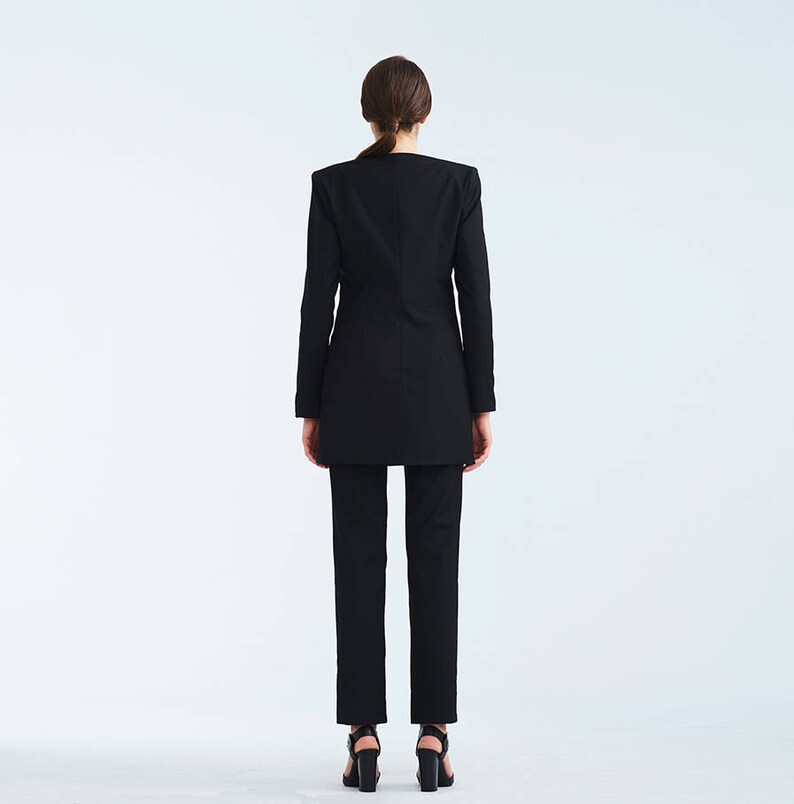 May include: A black blazer with a long, fitted silhouette. The blazer has a notched collar and long sleeves. The model is wearing black pants and black heels.