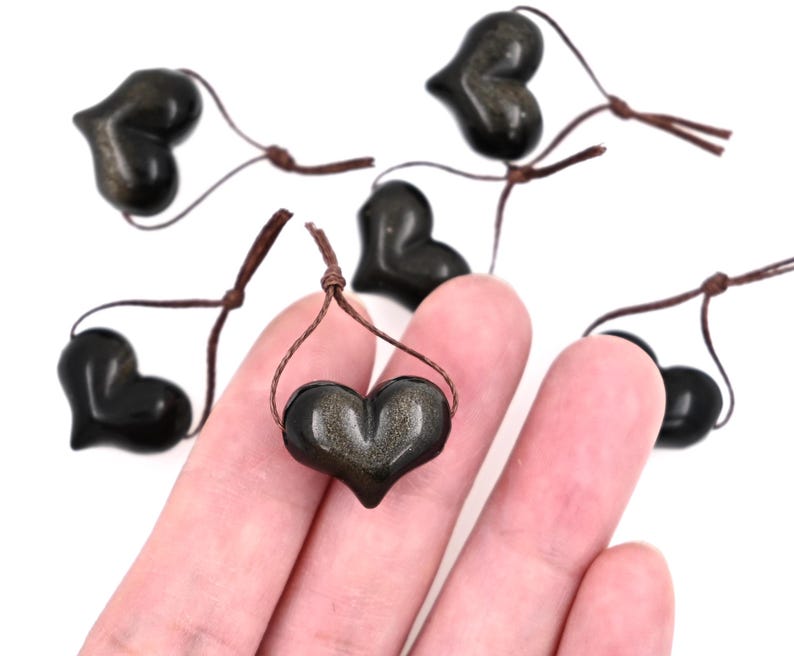 Puede incluir: Seis colgantes de coraz&oacute;n negro con motas doradas, cada uno en un cord&oacute;n marr&oacute;n.