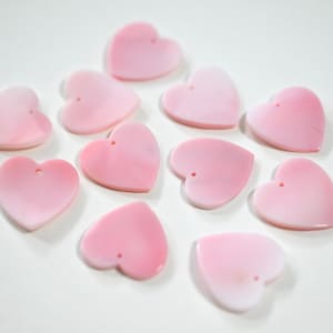May include: A selection of pale pink heart-shaped beads, each with a small hole at the top. The beads have a shiny surface and are arranged on a white background. Suitable for jewellery making and craft projects.