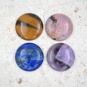 Könnte beinhalten: Vier runde, polierte Edelsteinkabochons in verschiedenen Farben. Die Cabochons sind in einem quadratischen Muster auf einem hellgrauen Hintergrund angeordnet. Die Cabochons sind hellbraun mit dunkelbraunen Streifen, hellrosa mit dunkelgrauen Adern, dunkelblau mit goldenen Flecken und helllila mit einer dunkelgrauen Ader.