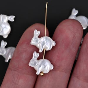 Może przedstawiać: Białe koraliki porcelanowe w kształcie królika z gładkim, błyszczącym wykończeniem. Koraliki są nawleczone na złoty drut.