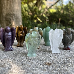 Könnte beinhalten: Sieben kleine Engelsfiguren, die aus verschiedenen Edelsteinen geschnitzt sind. Die Engel sind in einer Reihe auf einer grauen Steinoberfläche angeordnet. Die Engel sind lila, braun, grün, braun und weiß, grün und grau.
