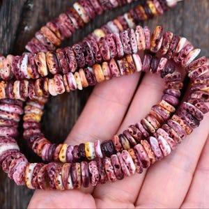 May include: A strand of circular, disc-shaped beads featuring a porous, textured surface. The beads display a variegated color palette of deep purple, magenta, cream, and orange tones, arranged in a continuous sequence on a thin string.