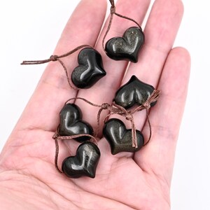 Puede incluir: Seis piedras negras en forma de coraz&oacute;n con motas doradas, enhebradas en cordones marrones. Las piedras est&aacute;n dispuestas en una mano, con los cordones atados juntos.