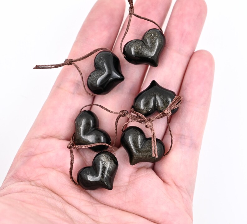 Puede incluir: Seis piedras negras en forma de coraz&oacute;n con motas doradas, enhebradas en cordones marrones. Las piedras est&aacute;n dispuestas en una mano, con los cordones atados juntos.