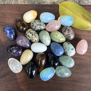 Op de afbeelding: Een verzameling gepolijste edelsteen eieren in verschillende kleuren, waaronder blauw, roze, groen en wit. De eieren zijn gerangschikt op een donker houten oppervlak, met een geel blad op de achtergrond. De edelstenen hebben verschillende patronen en texturen.