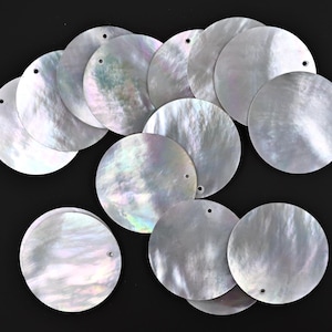 Puede incluir: Una colección de 14 discos redondos de concha blanca iridiscente con un agujero en el centro. Los discos están dispuestos en un patrón aleatorio.
