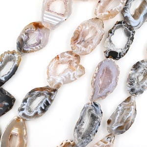 May include: A collection of irregular, oval-shaped agate slices arranged in strands. Each piece features a translucent, crystalline center surrounded by concentric bands of gray, brown, white, and black, with natural jagged edges and hollow centers.