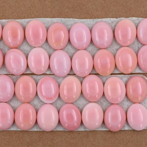 May include: A selection of oval, polished pink gemstones displayed on a white surface. The gemstones range in shades of pink, from pale to rose, and are set against a neutral brown backdrop.