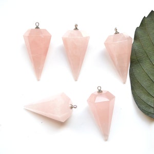 Może przedstawiać: Pięć fasetowanych wahadeł z różowego kwarcu ze srebrnymi metalowymi pętlami. Wahadła mają delikatny różowy kolor i są ułożone na białej powierzchni z zielonym liściem w tle.