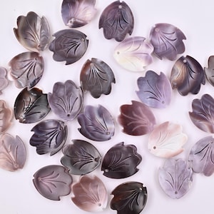 May include: A collection of iridescent, leaf-shaped pendants in shades of purple, gray, and white. Each pendant features a detailed vein design and a small hole for stringing. The pendants are scattered across a white surface.