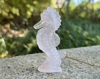 May include: A small, translucent pink seahorse figurine carved from rose quartz. The figurine is detailed with fins and a curled tail.