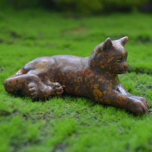 Puede incluir: Una figurita de cerámica de un gato con un patrón moteado marrón y negro, tumbada de lado con la cabeza girada hacia un lado. La figurita está descansando sobre un lecho de musgo verde.