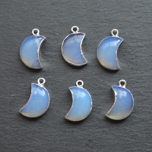 May include: Six crescent moon charms with silver-coloured edging and a pale blue, opalescent centre. Each charm has a small loop at the top for attaching to jewellery. The charms are arranged on a dark grey surface.