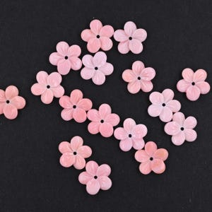 May include: A collection of fifteen small, five-petaled flower-shaped beads in a soft, light pink color. Each individual bead features a smooth, rounded petal design and a single, centered circular hole for threading or attachment purposes.
