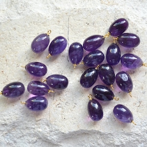 Puede incluir: Una colección de 18 cuentas de piedra preciosa de amatista con anillas de salto doradas. Las cuentas son de forma ovalada y tienen un color morado intenso.