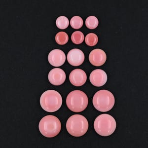 May include: A collection of eighteen smooth, round, pink cabochons arranged in six rows of three. The cabochons vary in size, with the smallest at the top and the largest at the bottom, all featuring a polished, glossy finish and a uniform pink hue.