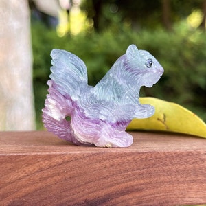Könnte beinhalten: Eine geschnitzte Fluoritsquirrel-Figur mit einem Farbspiel aus Lila, Grün und Weiß. Das Eichhörnchen sitzt auf einer Holzoberfläche.