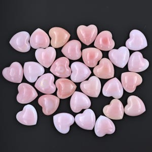 May include: A collection of pink heart-shaped shell cabochons. The cabochons are smooth and polished, with a natural, iridescent sheen.