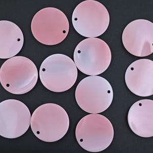 May include: A selection of pink, circular shell beads, each with a small hole near the edge. The beads have a pearlescent finish and range in colour from light pink to a deeper rose. The beads are displayed on a black background.