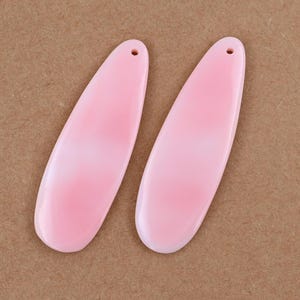 May include: Two pink, teardrop-shaped pendants with a hole at the top for stringing. The pendants have a gradient effect, transitioning from a darker pink at the bottom to a lighter pink at the top. The background is a solid brown.