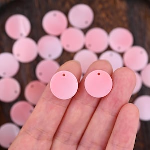 May include: A collection of small, circular, light pink flat discs, each featuring a single, centered hole near the top edge. These uniform, matte-finish components are arranged in a scattered pattern, with two pieces held up in the foreground.