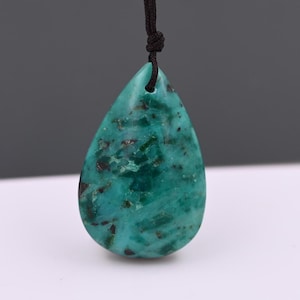 Puede incluir: Colgante en forma de lágrima con un patrón moteado en tonos turquesa y verde oscuro. Está suspendido de un cordón negro anudado en la parte superior. La piedra tiene una superficie pulida.