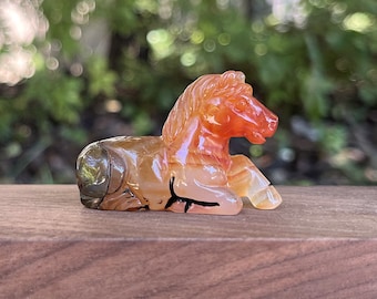 May include: A carved carnelian agate horse figurine. The horse is lying down with its head turned to the side. The agate is a mix of orange, brown, and white.