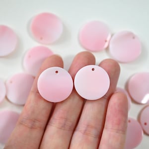 May include: Pale pink, circular charms with a small hole at the top. Several charms are scattered on a white surface, with two held in a hand. The charms have a smooth, glossy finish, suitable for jewellery making or crafting projects.