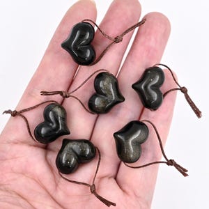 Puede incluir: Siete colgantes de piedra de sol en forma de coraz&oacute;n negro con cordones marrones. Los colgantes est&aacute;n dispuestos en una mano.