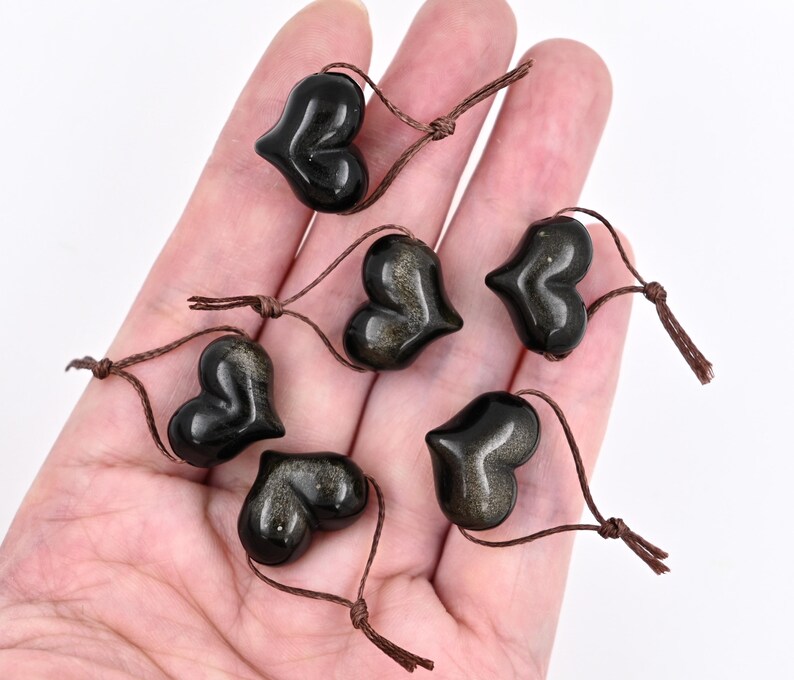Puede incluir: Siete colgantes de piedra de sol en forma de coraz&oacute;n negro con cordones marrones. Los colgantes est&aacute;n dispuestos en una mano.