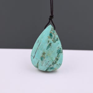 Puede incluir: Un colgante en forma de lágrima de piedra de color turquesa, suspendido de un cordón marrón oscuro. La piedra tiene un patrón moteado de inclusiones verdes oscuras y marrones, con finas líneas oscuras. El colgante es una piedra natural, para joyería.
