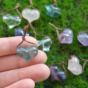 May include: A collection of heart-shaped crystal pendants, each with a different color, including clear, green, purple, and pink. The pendants are strung on brown cords and are displayed on a green mossy surface.