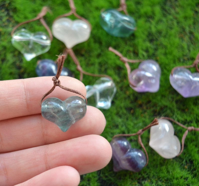 May include: A collection of heart-shaped crystal pendants, each with a different color, including clear, green, purple, and pink. The pendants are strung on brown cords and are displayed on a green mossy surface.