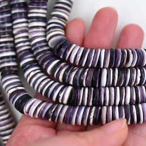 May include: Close-up of strands of purple and white shell beads. The beads are flat, disc-shaped, and strung together. The colour variation includes shades of purple, white, and black. The beads are held in a hand, showcasing their texture and arrangement.
