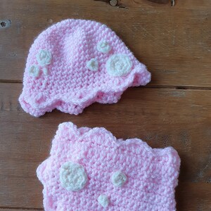 May include: A handmade pink crochet baby hat and matching bib set. The hat and bib feature white floral accents. The set is made of soft yarn and is perfect for a baby shower gift or newborn photo prop.