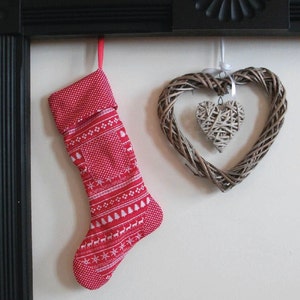 Puede incluir: Un calcetín navideño rojo con un patrón blanco de árboles, renos y copos de nieve cuelga de una cinta roja. También se muestra una corona en forma de corazón tejida con un corazón más pequeño en el interior, creando una escena festiva.