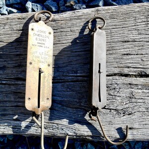 May include: Two antique brass hanging scales with a hook at the bottom. The scale on the left has a dial with markings from 0 to 50. The scale on the right has a plain dial. The scales are on a wooden surface. The scale on the left has the text "JOHN CHATILLON & SONS MAKERS NEW YORK U.S.A. NOT LEGAL FOR USE IN TRADE" printed on it.