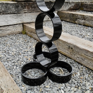 Puede incluir: Una escultura moderna para exteriores compuesta por anillos negros brillantes interconectados. La escultura se encuentra sobre un lecho de peque&ntilde;as piedras grises, con vigas de madera envejecida en el fondo, creando un contraste de texturas y colores.