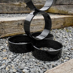 Puede incluir: Escultura de metal negro con formas circulares interconectadas. La escultura tiene un acabado brillante y est&aacute; colocada sobre un lecho de grava gris, con tablones de madera en el fondo. El dise&ntilde;o es moderno y abstracto.