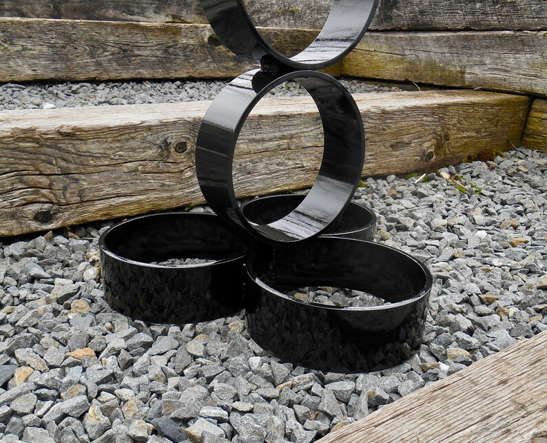 Puede incluir: Escultura de metal negro con formas circulares interconectadas. La escultura tiene un acabado brillante y est&aacute; colocada sobre un lecho de grava gris, con tablones de madera en el fondo. El dise&ntilde;o es moderno y abstracto.